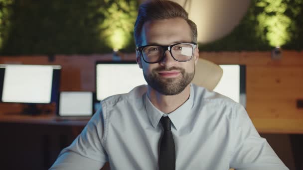 Joven feliz mirando a la cámara y sonriendo mientras se queda hasta tarde en la oficina — Vídeos de Stock