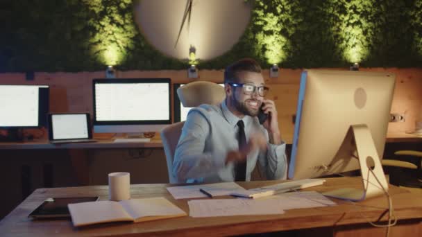 Щасливий молодий чоловік розмовляє по мобільному телефону, перебуваючи пізно в офісі — стокове відео