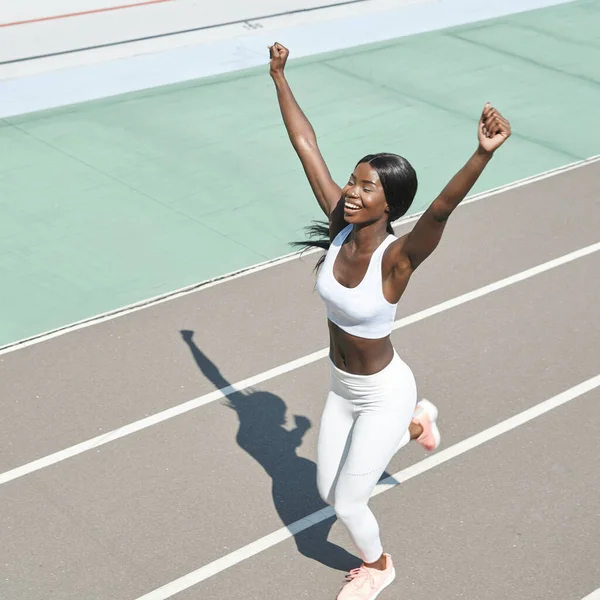 Вид сверху счастливой молодой африканской женщины в спортивной одежде, бегущей по дорожке и держащей руки поднятыми — стоковое фото