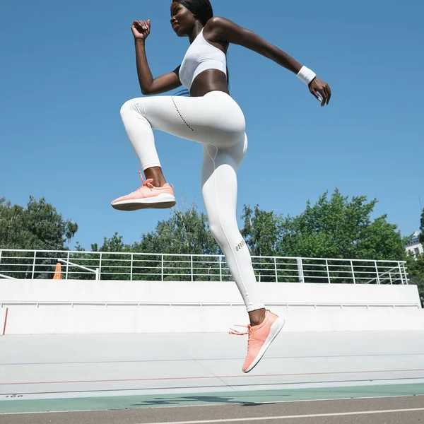 Mulher africana jovem confiante em roupas esportivas pulando na pista ao ar livre — Fotografia de Stock