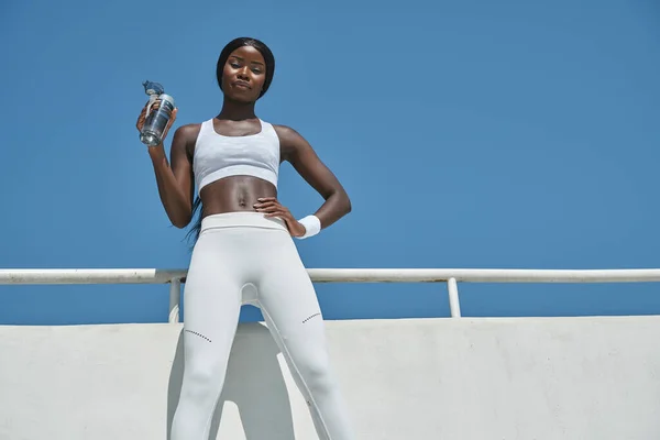 Vista de ángulo bajo de la hermosa joven africana en ropa deportiva sosteniendo botella con agua — Foto de Stock