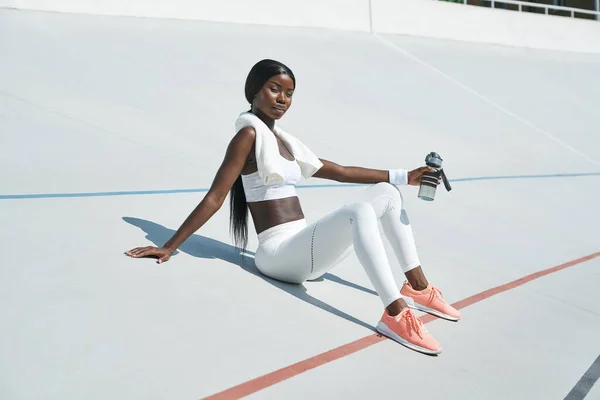 Hermosa joven africana en ropa deportiva sosteniendo botella con agua mientras está sentada en pista deportiva al aire libre —  Fotos de Stock