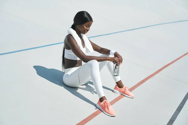 Hermosa joven africana en ropa deportiva sosteniendo botella con agua mientras está sentada en pista deportiva al aire libre —  Fotos de Stock