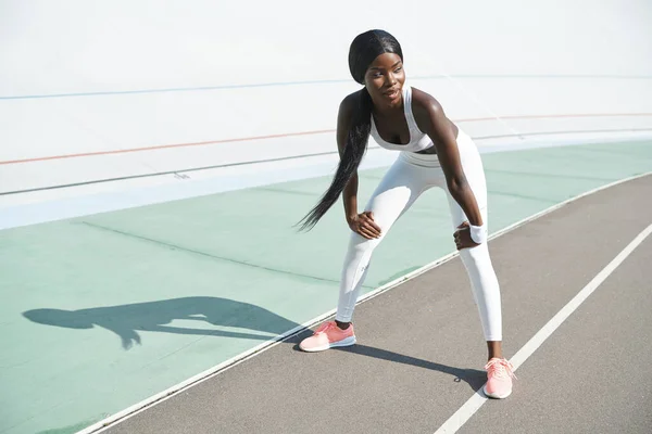 Comprimento total de bela jovem africana em roupas esportivas relaxante após o treinamento enquanto está de pé ao ar livre — Fotografia de Stock