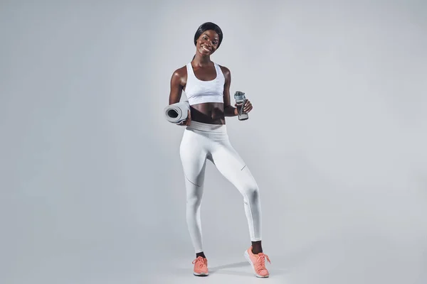 Счастливая молодая африканская женщина в спортивной одежде держит бутылку с водой и коврик для упражнений — стоковое фото