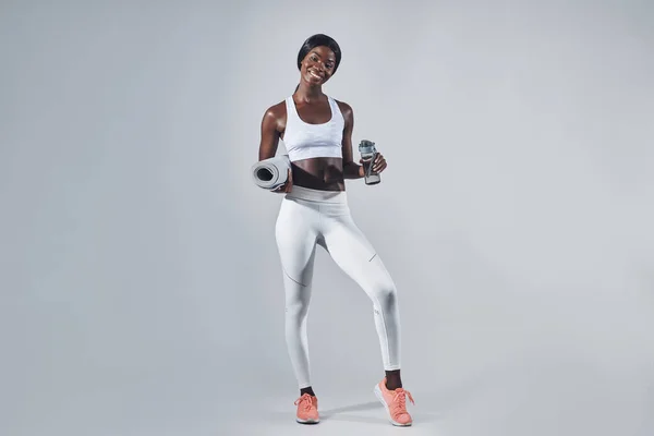 Happy young African woman in sports clothing holding bottle with water and exercise mat — Stock Photo, Image