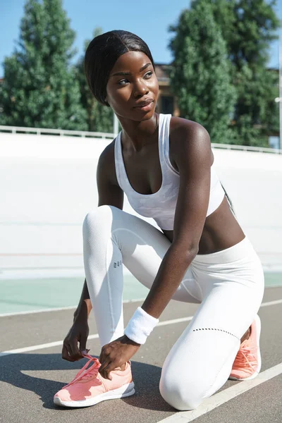 Selbstbewusste junge Afrikanerin in Sportkleidung bindet Schnürsenkel und schaut weg, während sie draußen auf der Laufbahn steht — Stockfoto