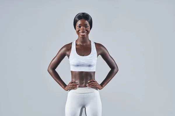 Mujer africana joven y segura en ropa deportiva cogida de la mano en la cadera mientras está de pie sobre un fondo gris —  Fotos de Stock