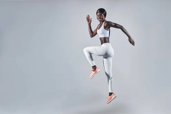 Longitud completa de mujer africana joven y confiada en ropa deportiva saltando sobre fondo gris — Foto de Stock