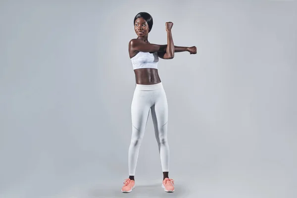 Fuld længde af smuk ung afrikansk kvinde i sportstøj strækker sig mod grå baggrund - Stock-foto