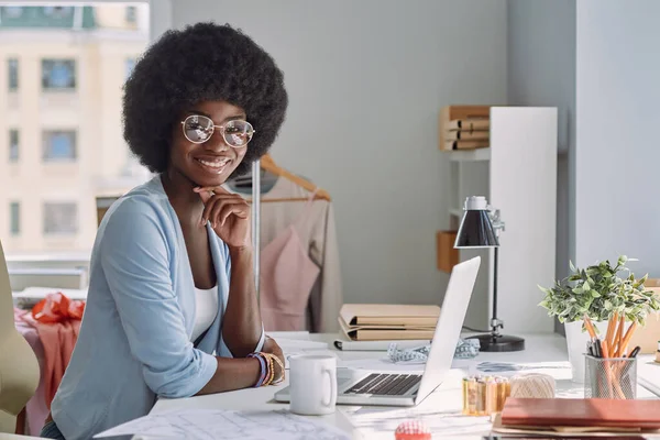 Hermosa joven africana mirando a la cámara y sonriendo mientras está sentada en su lugar de trabajo en el estudio de diseño —  Fotos de Stock