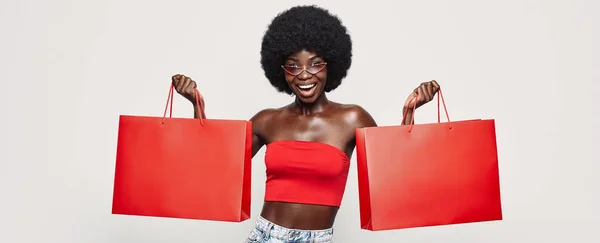 Mulher africana bonita carregando sacos de compras vermelhos e sorrindo enquanto está de pé contra fundo cinza — Fotografia de Stock