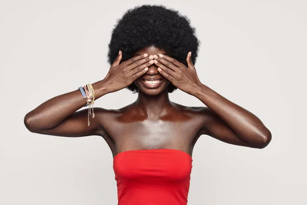Mulher africana bonita cobrindo os olhos com as mãos e sorrindo enquanto está de pé contra fundo cinza — Fotografia de Stock