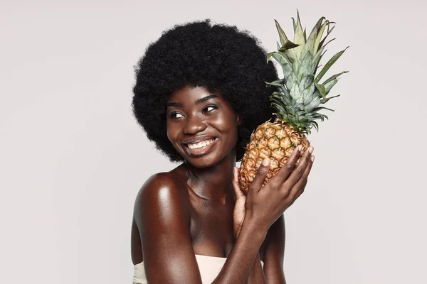 Retrato de una hermosa joven africana sosteniendo piña y sonriendo mientras está de pie sobre un fondo gris — Foto de Stock