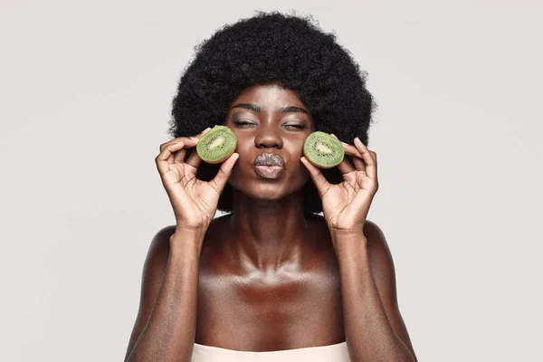 Retrato de una hermosa joven africana sosteniendo pedazos de kiwi cerca de la cara — Foto de Stock