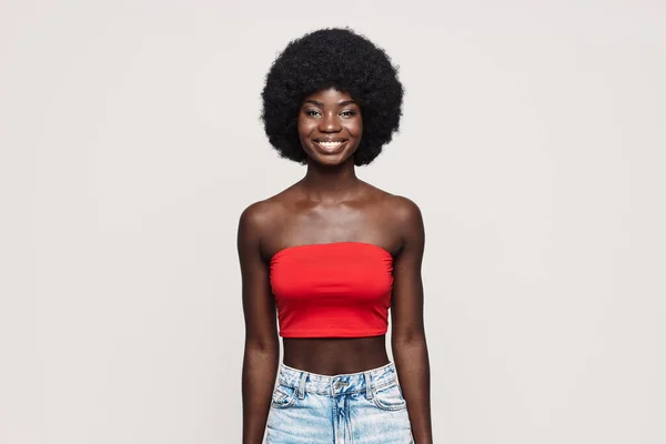Hermosa mujer africana joven mirando a la cámara y sonriendo mientras está de pie sobre un fondo gris —  Fotos de Stock