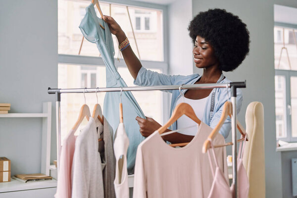 Красивая молодая африканская женщина осматривает платья и улыбается во время работы в дизайнерской студии