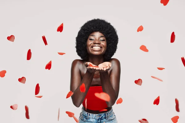 Beautiful young African woman looking at camera and blowing confetti while standing against gray background — Stock Photo, Image