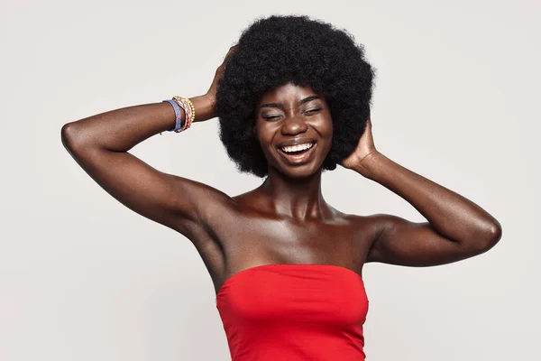 Hermosa joven africana cogida de la mano detrás de la cabeza y sonriendo mientras está de pie sobre un fondo gris — Foto de Stock