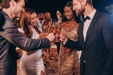 Formalite icabı giyinmiş bir grup insan şampanyayla kadeh kaldırıyor ve lüks partilerde vakit geçirirken gülümsüyor.