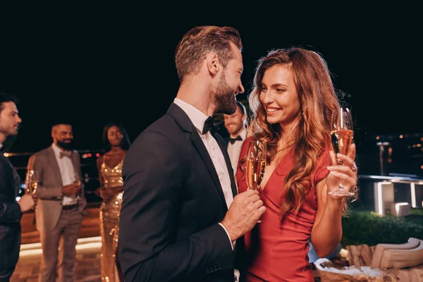 Hermosa pareja joven en ropa formal sosteniendo flautas de champán y sonriendo mientras pasa tiempo en una fiesta de lujo — Foto de Stock
