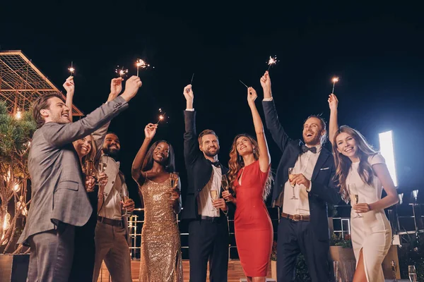 Gruppe schöner Menschen in Formalbekleidung, Wunderkerzen in der Hand und lächelnd, während sie Zeit auf einer Outdoor-Party verbringen — Stockfoto