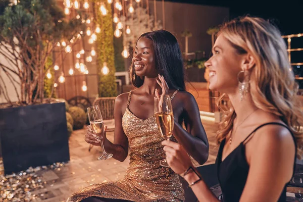 Duas mulheres bonitas em vestidos de noite bebendo champanhe e sorrindo ao passar o tempo em festa de luxo — Fotografia de Stock