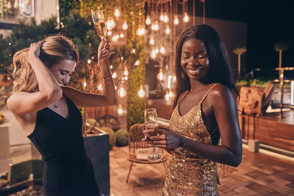 Duas mulheres bonitas em vestidos de noite dançando e sorrindo enquanto passa o tempo em festa de luxo — Fotografia de Stock