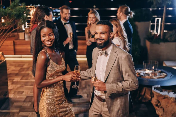 Hermosa joven pareja africana en ropa formal sosteniendo flautas de champán y sonriendo — Foto de Stock