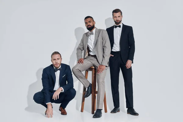 Three handsome young men in suits and bowties looking at camera while standing against gray background — Stock Photo, Image