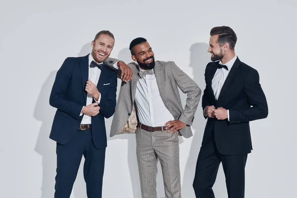 Three handsome young men in suits and bowties looking at camera and smiling while standing against gray background — Stock Photo, Image