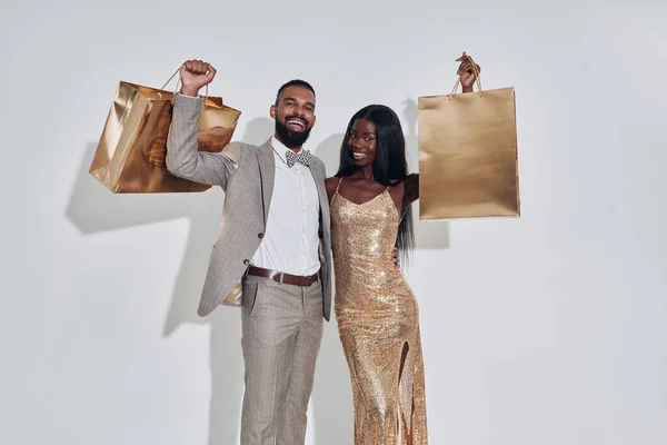 Hermosa pareja africana llevando bolsas de compras de color oro y sonriendo mientras está de pie sobre fondo gris — Foto de Stock