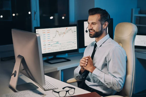 Szczęśliwy młody biznesmen w słuchawkach patrząc na komputer i uśmiechając się podczas pobytu do późna w biurze — Zdjęcie stockowe
