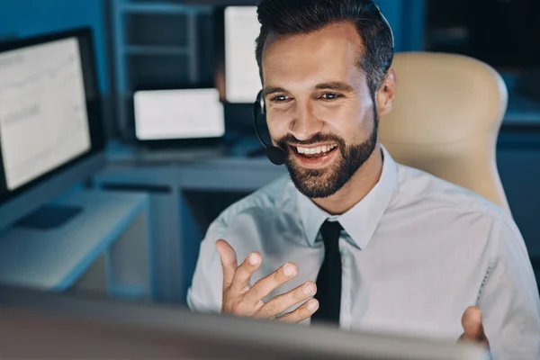 Joven feliz con auriculares mirando el ordenador y haciendo gestos mientras se queda hasta tarde en la oficina — Foto de Stock