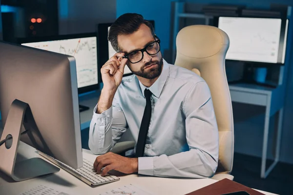 Впевнений молодий чоловік коригує окуляри, перебуваючи пізно в офісі — стокове фото