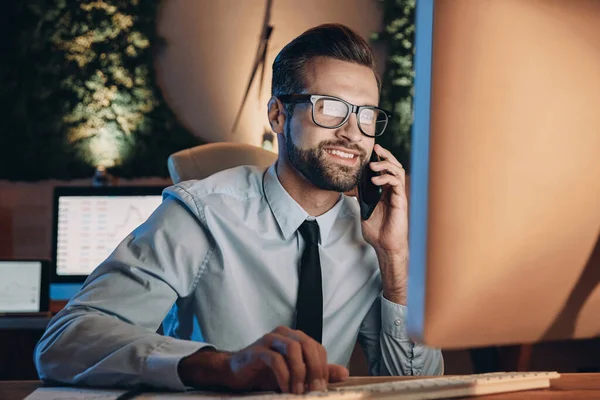 Cheerful young man working on computer and talking on mobile phone while staying late in the office — Stock Photo, Image