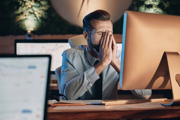 Frustrato giovane uomo che copre il viso con le mani durante il soggiorno fino a tardi in ufficio — Foto Stock