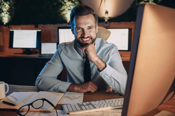 Jovem feliz olhando para a câmera e sorrindo enquanto fica até tarde no escritório — Fotografia de Stock