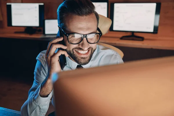 Jovem feliz falando no celular e olhando para o computador enquanto fica até tarde no escritório — Fotografia de Stock