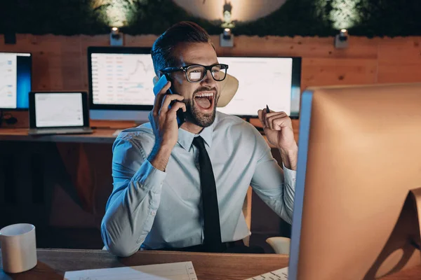 Szczęśliwy młody człowiek rozmawia przez telefon komórkowy i gestykuluje podczas pobytu do późna w biurze — Zdjęcie stockowe