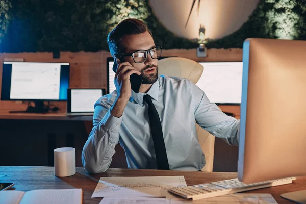 Jovem confiante trabalhando no computador e conversando no celular enquanto fica até tarde no escritório — Fotografia de Stock