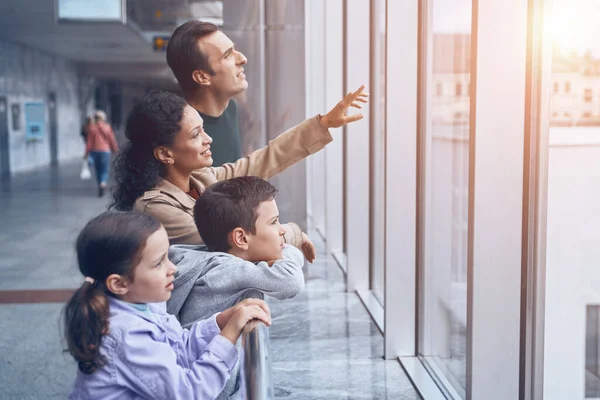 Familia feliz con dos niños pequeños mirando por una ventana mientras esperan su vuelo en la terminal del aeropuerto —  Fotos de Stock