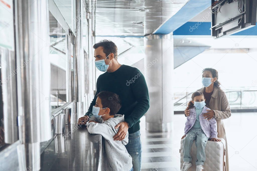 Family with two little kids in protective face masks buying tickets at ticket counter