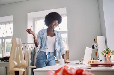 Güzel, genç Afrikalı bir kadın elinde kalemle iş yerinin yanında dururken mutlu görünüyor.