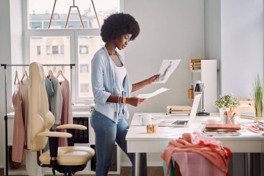 Güzel Afrikalı genç bir kadın ofisinde masasının yanında dururken moda tasarımı taslaklarını inceliyor.