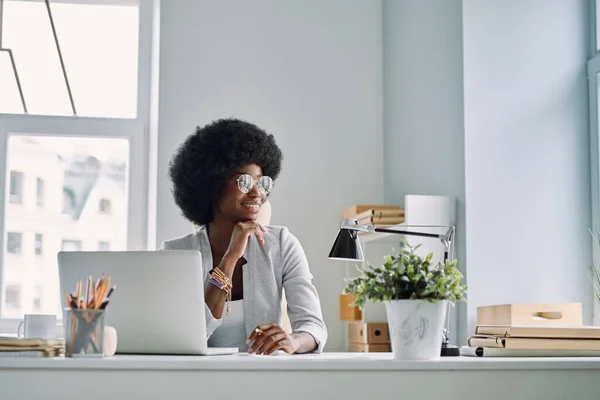 Belle jeune femme africaine tenant la main sur le menton et souriant tout en étant assise à son lieu de travail au bureau — Photo