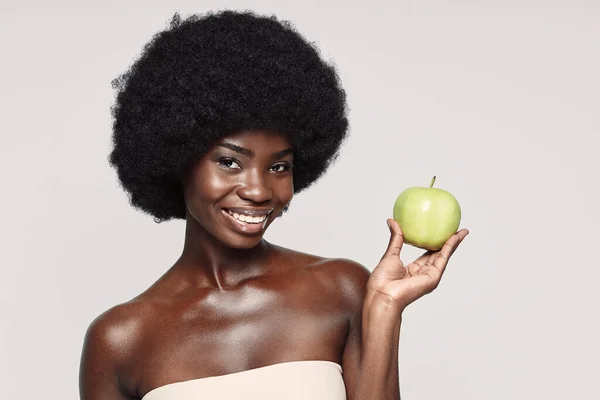 Gri arka planda dururken yeşil elma tutan ve gülümseyen güzel bir Afrikalı kadının portresi. — Stok fotoğraf