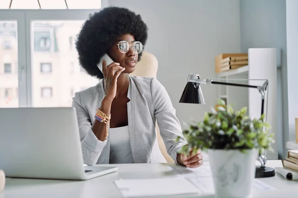 Красивая молодая африканская женщина разговаривает по мобильному телефону, сидя на рабочем месте в офисе — стоковое фото