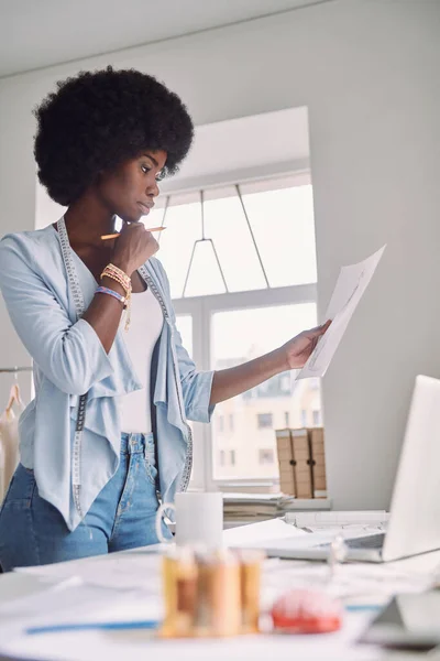 Krásná mladá africká žena analyzuje módní design skica, zatímco stojí v blízkosti svého stolu v kanceláři — Stock fotografie