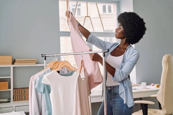 Hermosa joven africana examinando vestidos y sonriendo mientras trabaja en el estudio de diseño —  Fotos de Stock